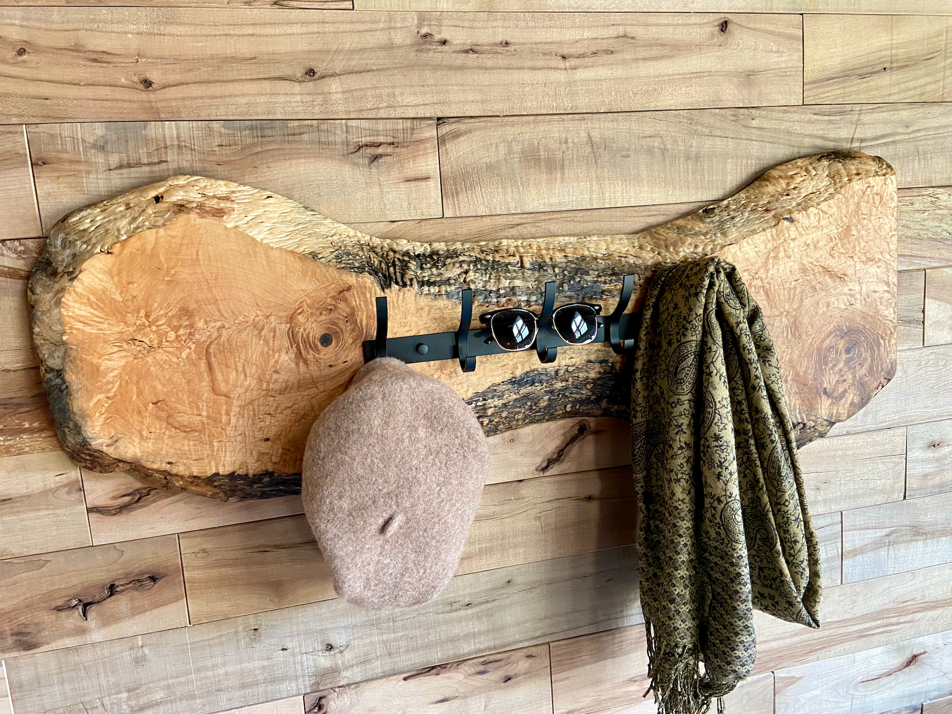 Live edge maple coat rack mounted on a wood-paneled wall with a hat and scarf hanging, featuring 5 matte black metal hooks and natural wood grain patterns.