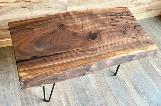 A handcrafted live edge walnut display table with black metal legs, placed against a couch. The live edge accent table showcases natural wood grain, knots, and organic edges, adding a rustic-modern touch to the space. A live edge walnut table styled with a candle, notebook, pen, a book, and a potted plant. The solid wood surface has a rich, natural finish, making it a perfect accent piece for an entryway or living space.