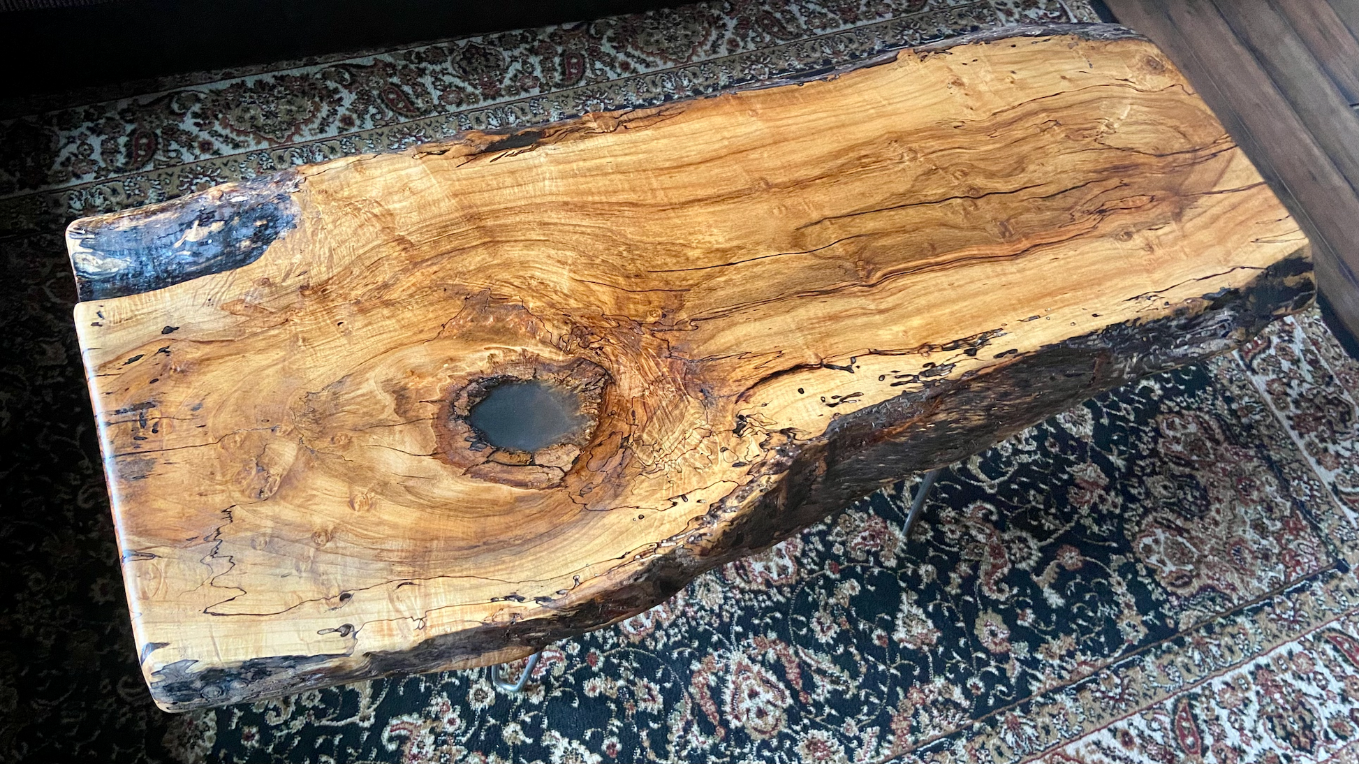 Live edge spalted ambrosia maple wood slab coffee table with a large knot on a patterned oriental rug.