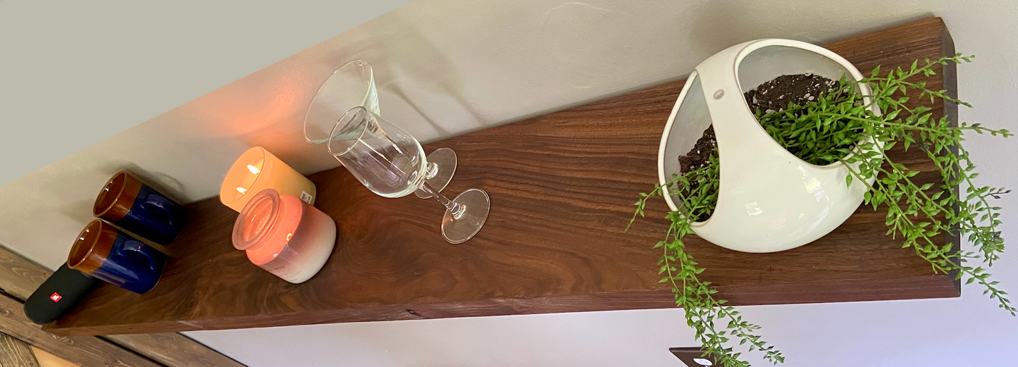 Front view of a handcrafted straight edge black walnut floating shelf mounted on a wall, displaying decorative items. Close-up of the solid black walnut wood grain, highlighting the shelf’s smooth finish. Side angle showcasing the shelf’s thickness and sturdy wall brackets.