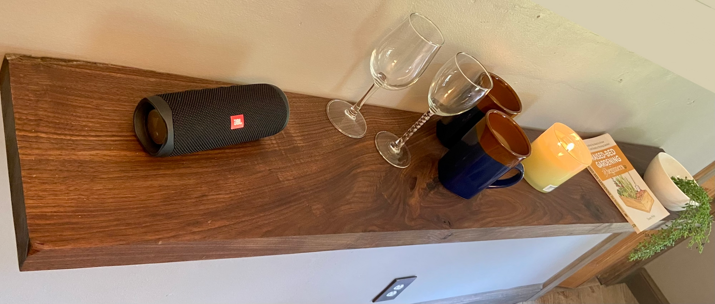 Front view of a handcrafted straight edge black walnut floating shelf mounted on a wall, displaying decorative items. Close-up of the solid black walnut wood grain, highlighting the shelf’s smooth finish. Side angle showcasing the shelf’s thickness and sturdy wall brackets.
