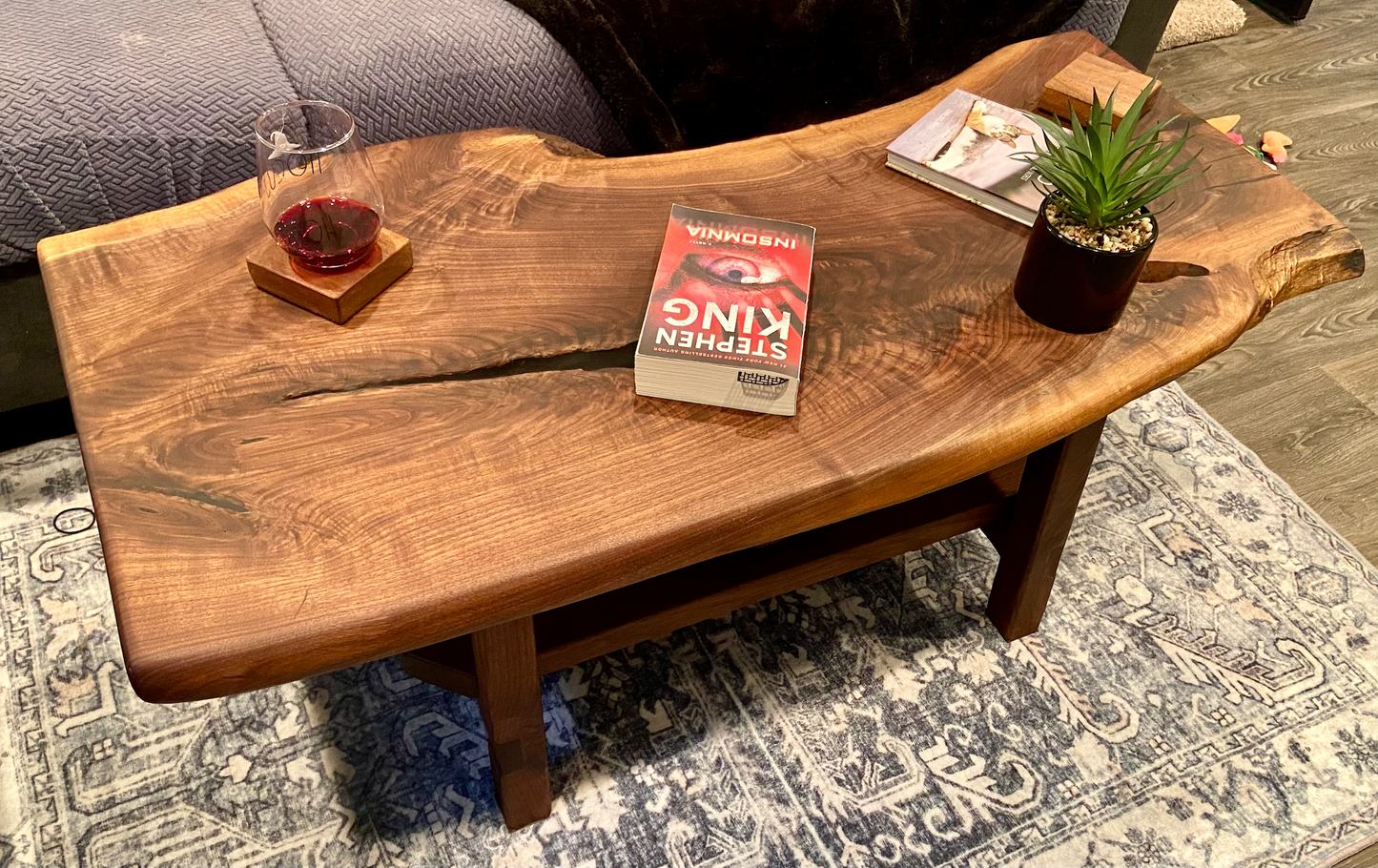 Live edge walnut wood slab coffee table with a book, glass of red wine, and small plant on a rug.